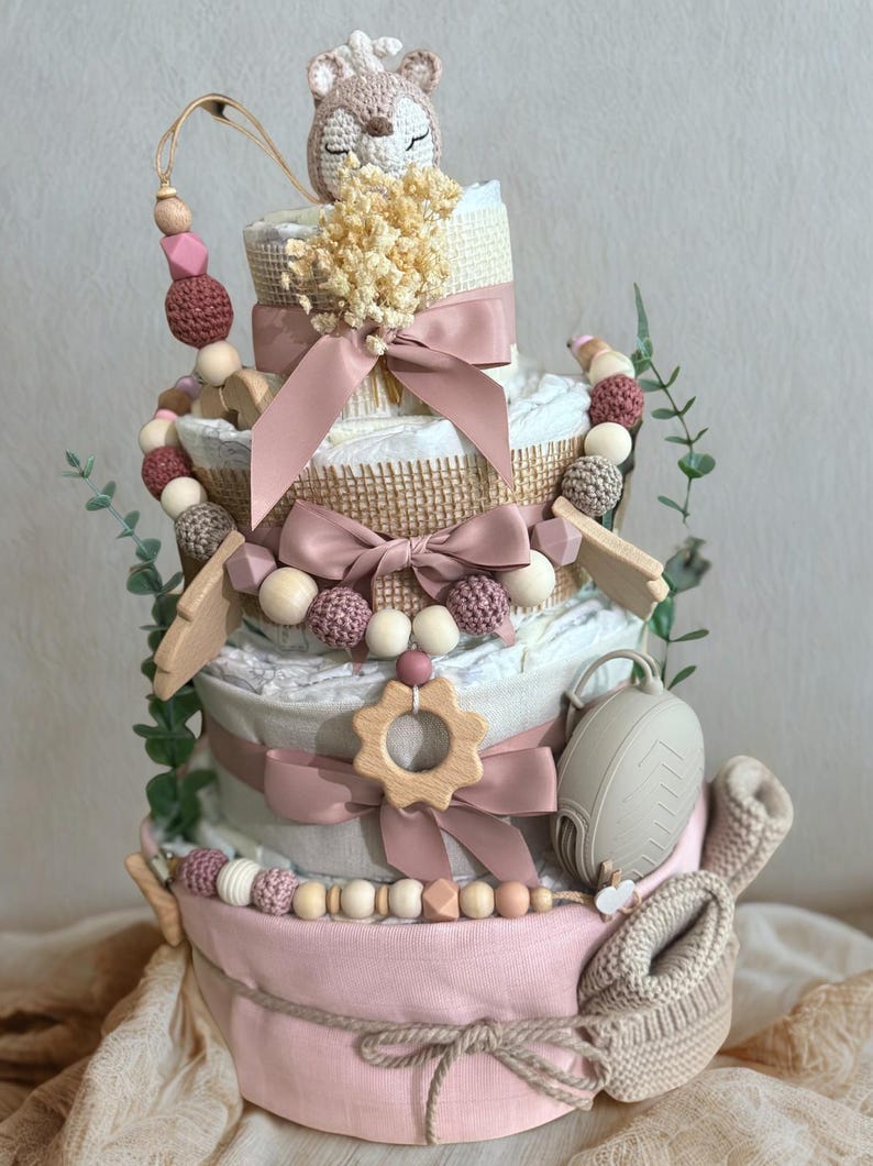 K&ouml;nnte beinhalten: Mehrst&ouml;ckige Windeltorte in Rosa, Wei&szlig; und Beige, verziert mit B&auml;ndern, Holzperlen und einem geh&auml;kelten Fuchs. Enth&auml;lt einen Schnuller, einen Bei&szlig;ring aus Holz und Babyschuhe. Ein einzigartiges Geschenk zur Babyparty.