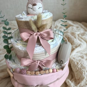 K&ouml;nnte beinhalten: Ein dreist&ouml;ckiger Windelkuchen in Rosa- und Beiget&ouml;nen, gekr&ouml;nt von einem geh&auml;kelten Reh. Der Kuchen ist mit rosa Schleifen, Holzperlen und dem Namen "NIHAN" verziert. Ein dekorativer Gegenstand ist an der Seite befestigt.