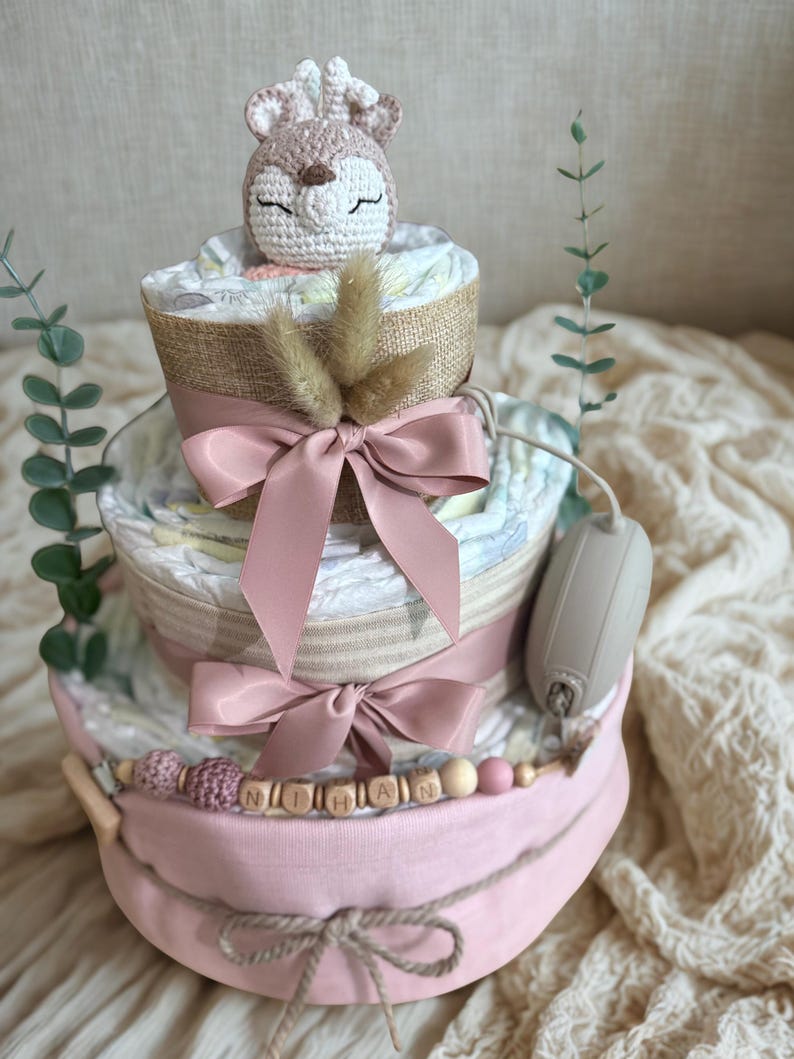 K&ouml;nnte beinhalten: Ein dreist&ouml;ckiger Windelkuchen in Rosa- und Beiget&ouml;nen, gekr&ouml;nt von einem geh&auml;kelten Reh. Der Kuchen ist mit rosa Schleifen, Holzperlen und dem Namen "NIHAN" verziert. Ein dekorativer Gegenstand ist an der Seite befestigt.