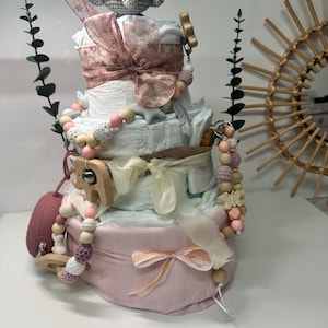 K&ouml;nnte beinhalten: Eine vierst&ouml;ckige Wickel-Torte, dekoriert mit rosa und wei&szlig;em Stoff, einem geh&auml;kelten grauen Elefanten, Holzperlen und einer Holzrassel. Die Torte ist von Gr&uuml;nzeug umgeben.