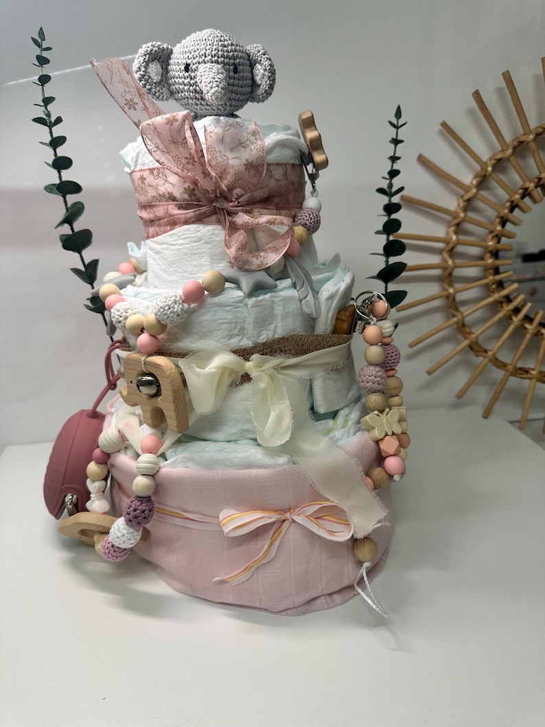 K&ouml;nnte beinhalten: Eine vierst&ouml;ckige Wickel-Torte, dekoriert mit rosa und wei&szlig;em Stoff, einem geh&auml;kelten grauen Elefanten, Holzperlen und einer Holzrassel. Die Torte ist von Gr&uuml;nzeug umgeben.