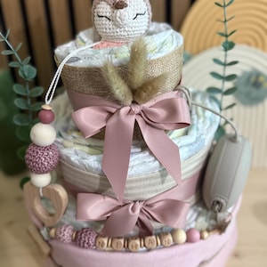 K&ouml;nnte beinhalten: Ein dreist&ouml;ckiger Babygeschenkkuchen mit einem geh&auml;kelten Hirschaufsatz. Der Kuchen ist mit rosa B&auml;ndern und einer Holzperlen-Girlande mit einem Namen verziert. Der Kuchen besteht aus Windeln und anderen Babyartikeln.