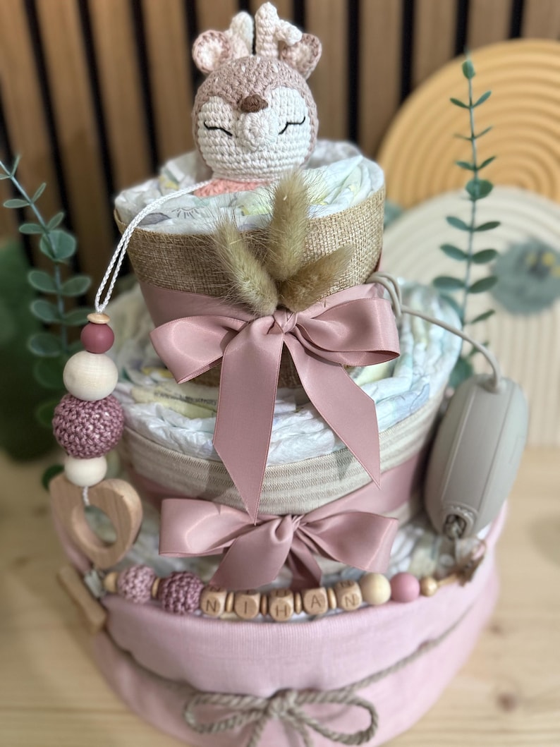 K&ouml;nnte beinhalten: Ein dreist&ouml;ckiger Babygeschenkkuchen mit einem geh&auml;kelten Hirschaufsatz. Der Kuchen ist mit rosa B&auml;ndern und einer Holzperlen-Girlande mit einem Namen verziert. Der Kuchen besteht aus Windeln und anderen Babyartikeln.