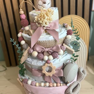 K&ouml;nnte beinhalten: Mehrst&ouml;ckige Babygeschenktorte in Rosa, Wei&szlig; und Beige. Die Torte ist mit B&auml;ndern, Holzperlen, einem geh&auml;kelten Reh und einem Schnuller verziert. Die Torte ist ein kreatives Geschenk zur Babyparty.