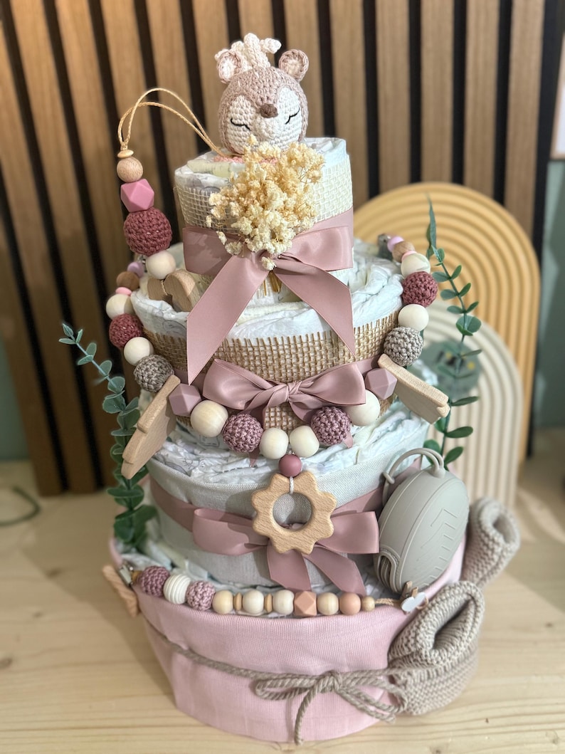 K&ouml;nnte beinhalten: Mehrst&ouml;ckige Babygeschenktorte in Rosa, Wei&szlig; und Beige. Die Torte ist mit B&auml;ndern, Holzperlen, einem geh&auml;kelten Reh und einem Schnuller verziert. Die Torte ist ein kreatives Geschenk zur Babyparty.