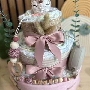 K&ouml;nnte beinhalten: Ein dreist&ouml;ckiger Babygeschenkkuchen mit einem geh&auml;kelten Hirschaufsatz. Der Kuchen ist mit rosa B&auml;ndern und einer Holzperlen-Girlande mit einem Namen verziert. Der Kuchen besteht aus Windeln und anderen Babyartikeln.