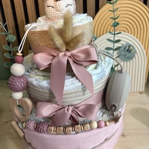 K&ouml;nnte beinhalten: Ein dreist&ouml;ckiger Windelkuchen mit einem geh&auml;kelten Hirschaufsatz, rosa Schleifen und dem Namen "NITHAN" in Holzperlen geschrieben. Der Kuchen ist mit Trockenblumen und einem Schnullerclip verziert.