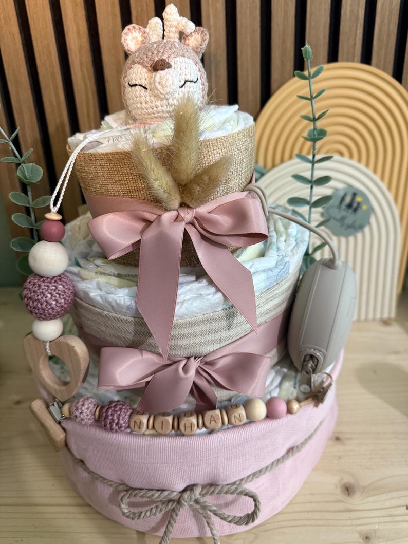 K&ouml;nnte beinhalten: Ein dreist&ouml;ckiger Windelkuchen mit einem geh&auml;kelten Hirschaufsatz, rosa Schleifen und dem Namen "NITHAN" in Holzperlen geschrieben. Der Kuchen ist mit Trockenblumen und einem Schnullerclip verziert.