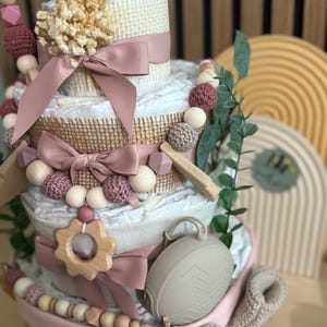 K&ouml;nnte beinhalten: Mehrst&ouml;ckige Windeltorte, verziert mit rosa B&auml;ndern, Holzperlen und einem geh&auml;kelten Eulenaufsatz. Der Kuchen enth&auml;lt einen Bei&szlig;ring aus Holz, einen grauen Schnullerhalter und dekoratives Gr&uuml;n. Ein Geschenk zur Babyparty.