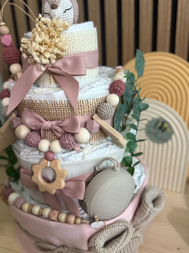 K&ouml;nnte beinhalten: Mehrst&ouml;ckige Windeltorte, verziert mit rosa B&auml;ndern, Holzperlen und einem geh&auml;kelten Eulenaufsatz. Der Kuchen enth&auml;lt einen Bei&szlig;ring aus Holz, einen grauen Schnullerhalter und dekoratives Gr&uuml;n. Ein Geschenk zur Babyparty.