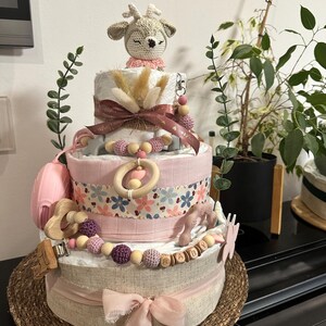 K&ouml;nnte beinhalten: Eine dreist&ouml;ckige Wickel-Torte, verziert mit einem geh&auml;kelten Reh, rosa und braunen B&auml;ndern, Holzperlen und einem Holz-Schild 'BABY'. Die Torte steht auf einer geflochtenen Matte.