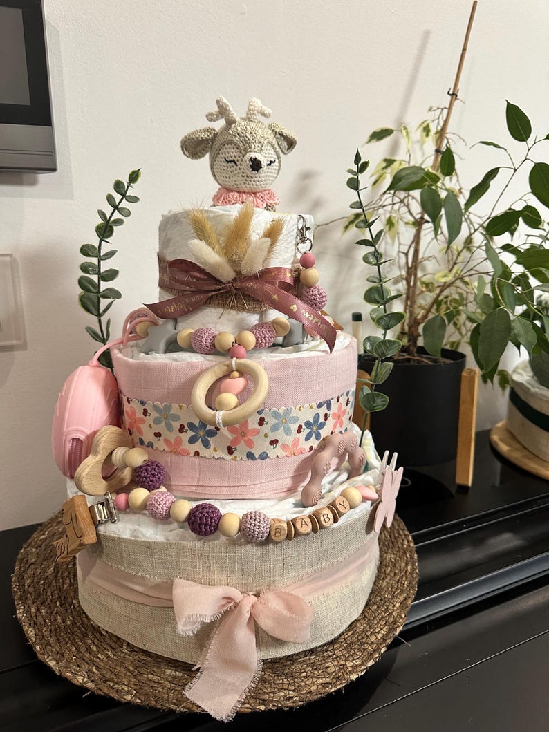 K&ouml;nnte beinhalten: Eine dreist&ouml;ckige Wickel-Torte, verziert mit einem geh&auml;kelten Reh, rosa und braunen B&auml;ndern, Holzperlen und einem Holz-Schild 'BABY'. Die Torte steht auf einer geflochtenen Matte.
