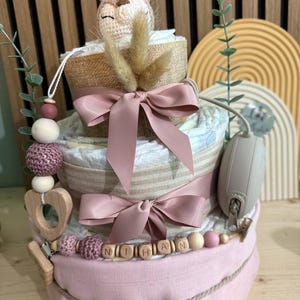 K&ouml;nnte beinhalten: Windeltorte mit drei Etagen, Giraffenaufsatz, rosa Schleifen und einer Holzperlenkette mit der Aufschrift "NITHAN". Der Kuchen ist mit Windeln, B&auml;ndern und einem Schnullerhalter verziert. Ein bezauberndes Geschenk zur Babyparty.