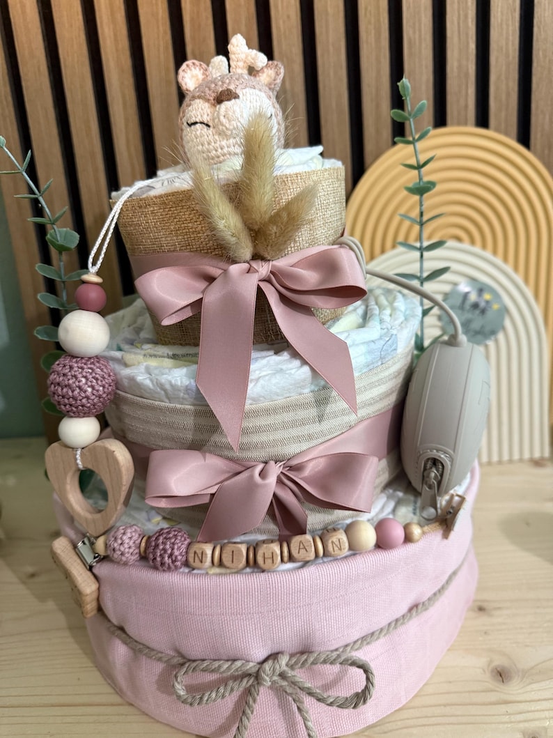 K&ouml;nnte beinhalten: Windeltorte mit drei Etagen, Giraffenaufsatz, rosa Schleifen und einer Holzperlenkette mit der Aufschrift "NITHAN". Der Kuchen ist mit Windeln, B&auml;ndern und einem Schnullerhalter verziert. Ein bezauberndes Geschenk zur Babyparty.