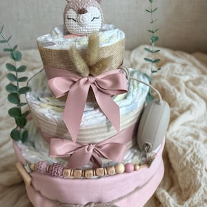 K&ouml;nnte beinhalten: Dreist&ouml;ckige Windeltorte in Rosa und Beige. Sie hat rosa Schleifen, Holzperlen und einen geh&auml;kelten Hirsch als Topper. Der Name "Nihan" ist in Holzbuchstaben geschrieben. Ein bezauberndes Babygeschenk.