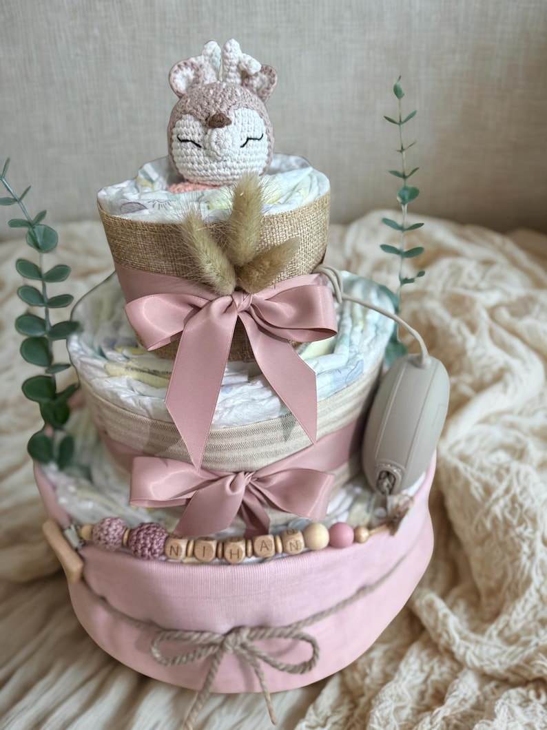 K&ouml;nnte beinhalten: Dreist&ouml;ckige Windeltorte in Rosa und Beige. Sie hat rosa Schleifen, Holzperlen und einen geh&auml;kelten Hirsch als Topper. Der Name "Nihan" ist in Holzbuchstaben geschrieben. Ein bezauberndes Babygeschenk.