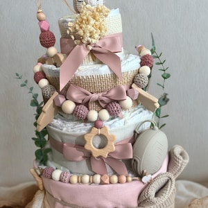 K&ouml;nnte beinhalten: Mehrst&ouml;ckige Windeltorte in Rosa, Beige und Wei&szlig;, verziert mit B&auml;ndern, Holzperlen und einem geh&auml;kelten Fuchs. Enth&auml;lt einen Schnuller, Holzformen und Babyschuhe. Ein einzigartiges Geschenk zur Babyparty.
