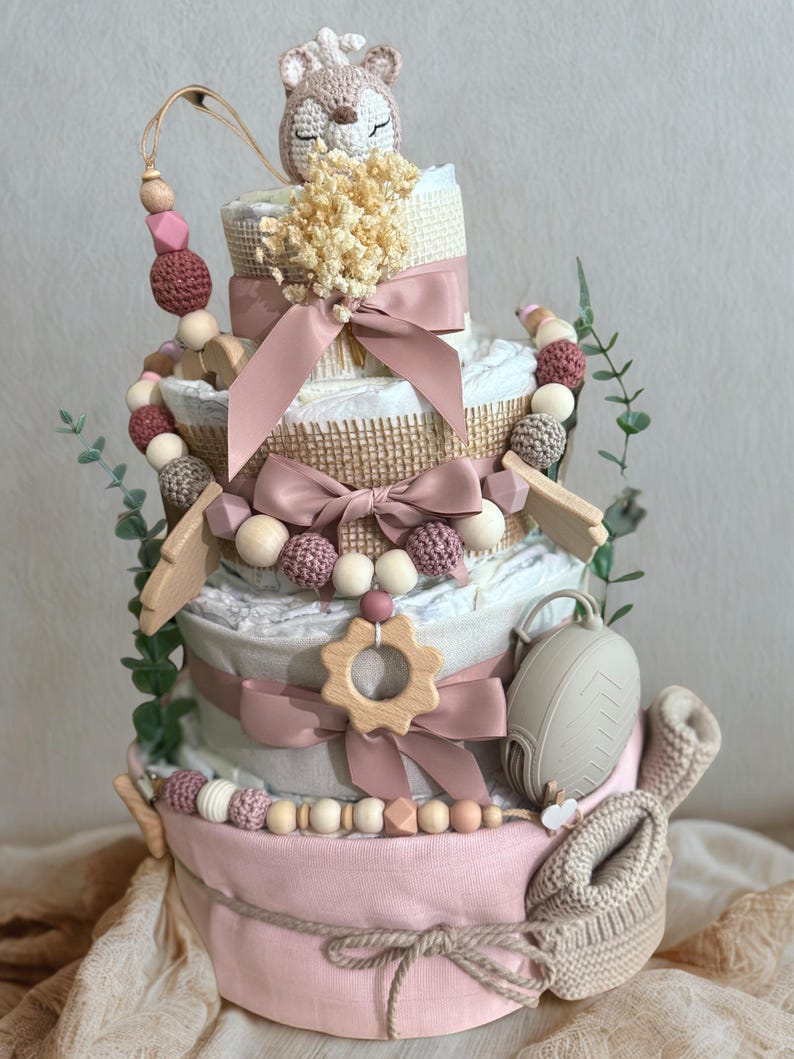 K&ouml;nnte beinhalten: Mehrst&ouml;ckige Windeltorte in Rosa, Beige und Wei&szlig;, verziert mit B&auml;ndern, Holzperlen und einem geh&auml;kelten Fuchs. Enth&auml;lt einen Schnuller, Holzformen und Babyschuhe. Ein einzigartiges Geschenk zur Babyparty.