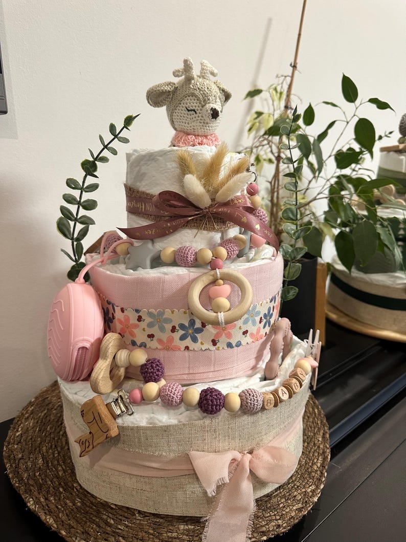 K&ouml;nnte beinhalten: Eine dreist&ouml;ckige Windeltorte, verziert mit einem geh&auml;kelten Reh, Holzperlen und einem rosa-wei&szlig;en Band. Die Torte ist mit einem Blumenmuster verziert und das Wort "BABY" ist in Holzbuchstaben geschrieben.