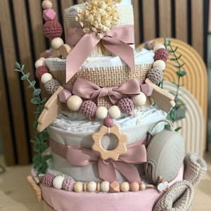 K&ouml;nnte beinhalten: Ein mehrst&ouml;ckiger Babygeschenkkuchen mit rosa Basis und Windelschichten, verziert mit B&auml;ndern, Holzperlen und einem geh&auml;kelten Rehkitz-Topper. Enth&auml;lt einen Schnuller und ein Paar Babyschuhe.