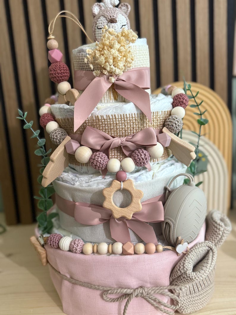 K&ouml;nnte beinhalten: Ein mehrst&ouml;ckiger Babygeschenkkuchen mit rosa Basis und Windelschichten, verziert mit B&auml;ndern, Holzperlen und einem geh&auml;kelten Rehkitz-Topper. Enth&auml;lt einen Schnuller und ein Paar Babyschuhe.