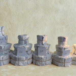 K&ouml;nnte beinhalten: F&uuml;nf Stapel Windeln, jeweils verpackt und mit Bindfaden verschn&uuml;rt, werden gezeigt. Jeder Stapel hat ein Etikett mit einer Gr&ouml;&szlig;enangabe: XXL 53, 44, 35, 21 und Mini 15. Die Windeln sind hellgrau mit einem dezenten Blumenmuster.