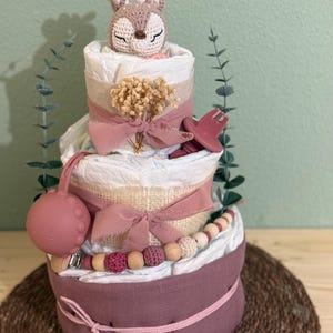 K&ouml;nnte beinhalten: Eine dreist&ouml;ckige Wickel-Torte, verziert mit einem geh&auml;kelten Hirsch-Spielzeug, rosa und beige B&auml;ndern, Holzperlen und einem rosa Silikon-Bei&szlig;ring. Die Torte steht auf einer braunen gewebten Matte.