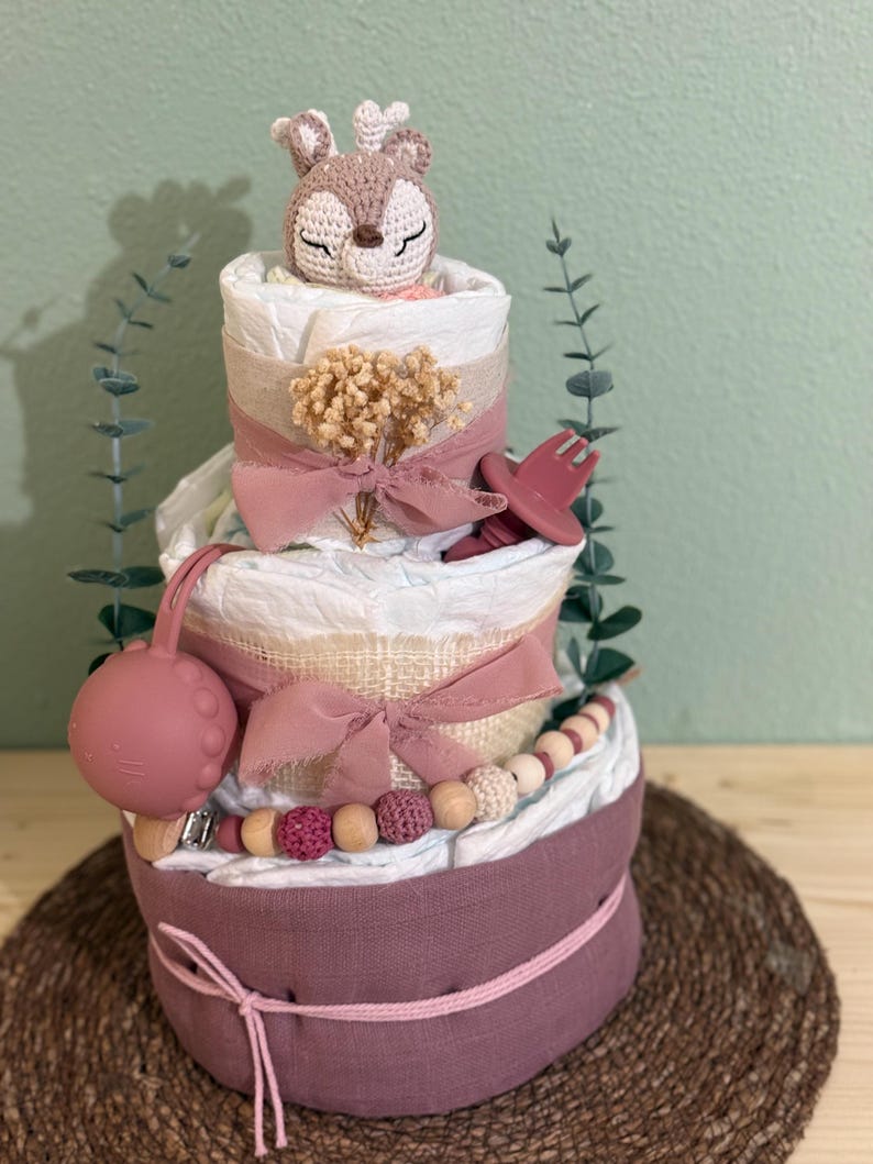 K&ouml;nnte beinhalten: Eine dreist&ouml;ckige Wickel-Torte, verziert mit einem geh&auml;kelten Hirsch-Spielzeug, rosa und beige B&auml;ndern, Holzperlen und einem rosa Silikon-Bei&szlig;ring. Die Torte steht auf einer braunen gewebten Matte.