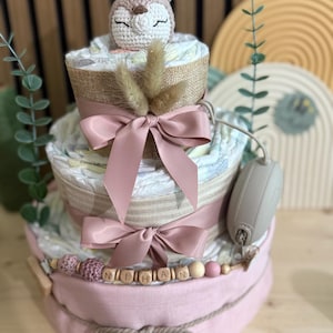 K&ouml;nnte beinhalten: Dreist&ouml;ckige Windeltorte mit geh&auml;keltem Hirschaufsatz. Der Kuchen hat rosa B&auml;nder, eine mit Jute umwickelte Schicht und eine Holzperlen-Girlande mit der Aufschrift "NIHAN". Ein Schnuller ist befestigt. Ein Geschenk zur Babyparty.