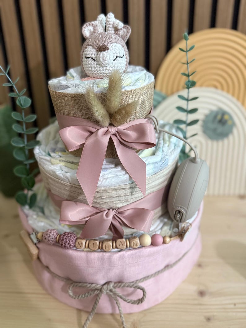 K&ouml;nnte beinhalten: Dreist&ouml;ckige Windeltorte mit geh&auml;keltem Hirschaufsatz. Der Kuchen hat rosa B&auml;nder, eine mit Jute umwickelte Schicht und eine Holzperlen-Girlande mit der Aufschrift "NIHAN". Ein Schnuller ist befestigt. Ein Geschenk zur Babyparty.