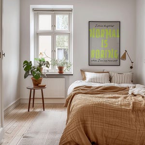 May include: A bedroom with a bed covered in a tan textured blanket and striped pillows. A framed poster with the words "NORMAL IS BORING" hangs above the bed. A small wooden table holds a potted plant, and a window is visible in the background.