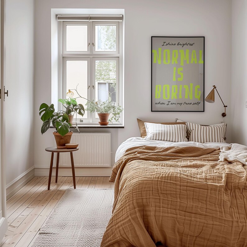 May include: A bedroom with a bed covered in a tan textured blanket and striped pillows. A framed poster with the words "NORMAL IS BORING" hangs above the bed. A small wooden table holds a potted plant, and a window is visible in the background.
