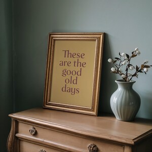 May include: A framed print with the text "These are the good old days" in a gold frame. The print is propped on a wooden dresser, next to a vase with white flowers. The background is a muted teal color.