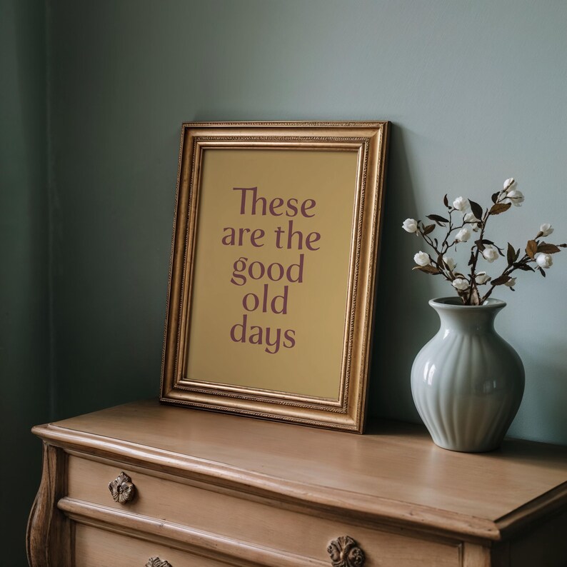 May include: A framed print with the text "These are the good old days" in a gold frame. The print is propped on a wooden dresser, next to a vase with white flowers. The background is a muted teal color.