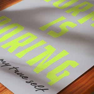 May include: A gray poster with the words "WORK IS WORKING" in bright neon yellow block letters. The phrase "my true self" is written in black cursive at the bottom. The poster is on a wooden surface.