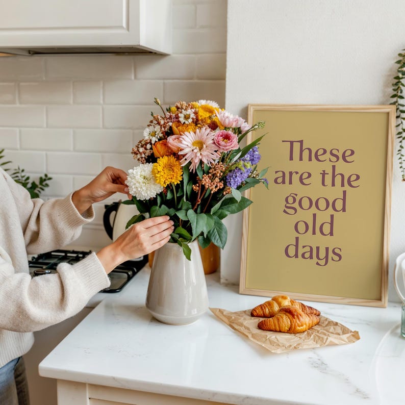 May include: A bouquet of colorful flowers in a gray vase on a white countertop. A framed print with the text "These are the good old days" is next to the vase. Croissants rest on a piece of brown paper.