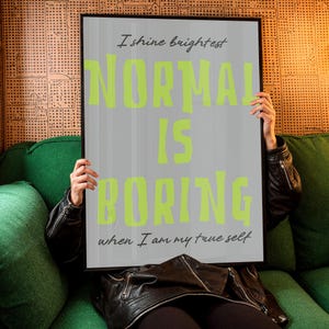 May include: A framed poster with the text "I shine brightest. NORMAL IS BORING when I am my true self" in lime green and gray. The poster is held by a person wearing a black leather jacket, sitting on a green couch.