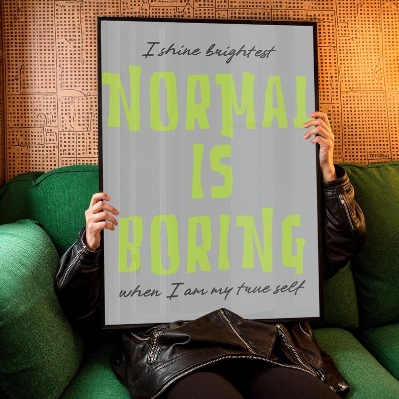 May include: A framed poster with the text "I shine brightest. NORMAL IS BORING when I am my true self" in lime green and gray. The poster is held by a person wearing a black leather jacket, sitting on a green couch.