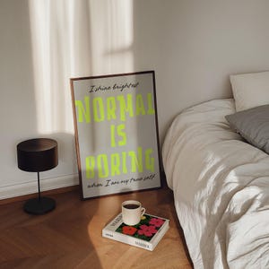 May include: A framed poster with the text "NORMAL IS BORING" in neon green, leaning against a wall. A black lamp and a book with a coffee cup on top are on the wooden floor. The book has red and pink flowers on the cover.