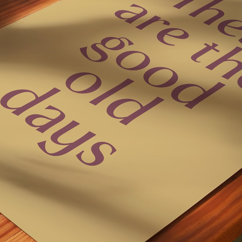 May include: A beige poster with the phrase "the good old days" printed in a purple, serif font. The poster is angled, with a wooden surface visible at the bottom. The text is large and centered.