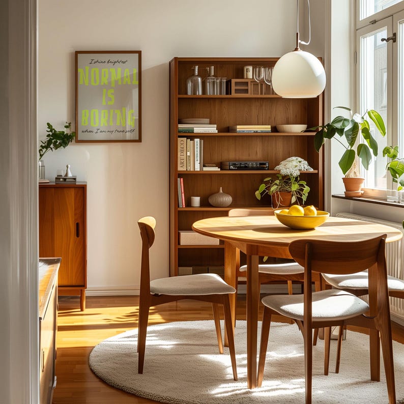 May include: A dining room scene featuring a round wooden table and chairs, a wooden bookshelf, and a framed poster with the text "NORMAL IS BORING." The room is bathed in natural light, with a rug and plants adding warmth.