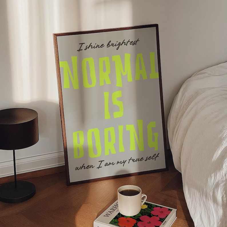 May include: A framed poster with the text "NORMAL IS BORING" in bright neon green, with the phrases "I shine brightest" and "when I am my true self". The poster is leaning against a wall next to a bed and a small table.