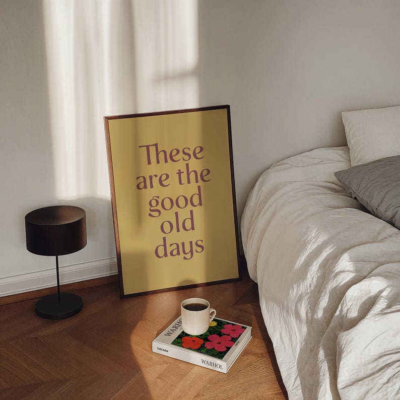 May include: A framed poster with the text "These are the good old days" in a brown font. A white coffee mug sits on a book with red and pink flowers. A black lamp and a bed with white linens are also in the image.