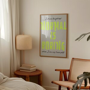 May include: A framed poster with the text "NORMAL IS BORING" in bright neon green on a gray background. The poster is in a room with a lamp, a small table, and a wooden chair.
