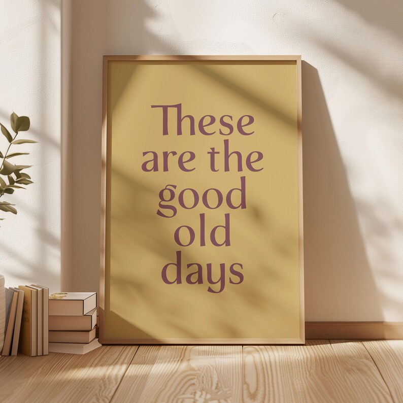 May include: A framed print with the text "These are the good old days" in a purple font on a yellow background. The frame is a light wood color. The print is leaning against a white wall, with a stack of books and a plant visible.
