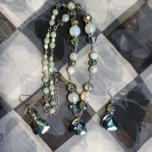 May include: A silver necklace with green and white beads and a matching pair of earrings with teardrop-shaped crystals. The necklace has a silver chain and a teardrop-shaped crystal pendant.