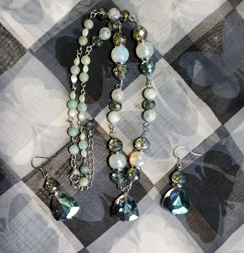 May include: A silver necklace with green and white beads and a matching pair of earrings with teardrop-shaped crystals. The necklace has a silver chain and a teardrop-shaped crystal pendant.