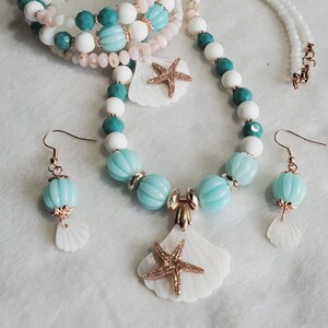 May include: A white and turquoise beaded necklace with a seashell pendant and starfish charm. The necklace is paired with matching earrings featuring seashells.