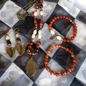 May include: A set of carnelian gemstone jewelry featuring a necklace, two bracelets, and earrings. The necklace has a leaf pendant and the bracelets have leaf charms. The jewelry is made with brown and orange carnelian beads and white beads.