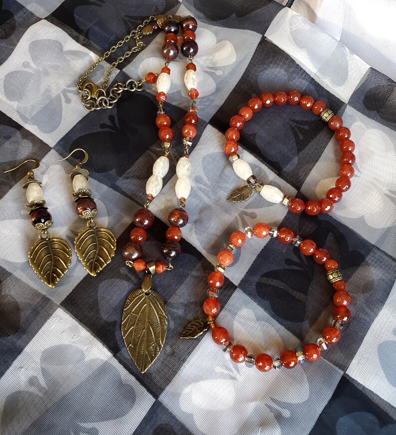May include: A set of carnelian gemstone jewelry featuring a necklace, two bracelets, and earrings. The necklace has a leaf pendant and the bracelets have leaf charms. The jewelry is made with brown and orange carnelian beads and white beads.